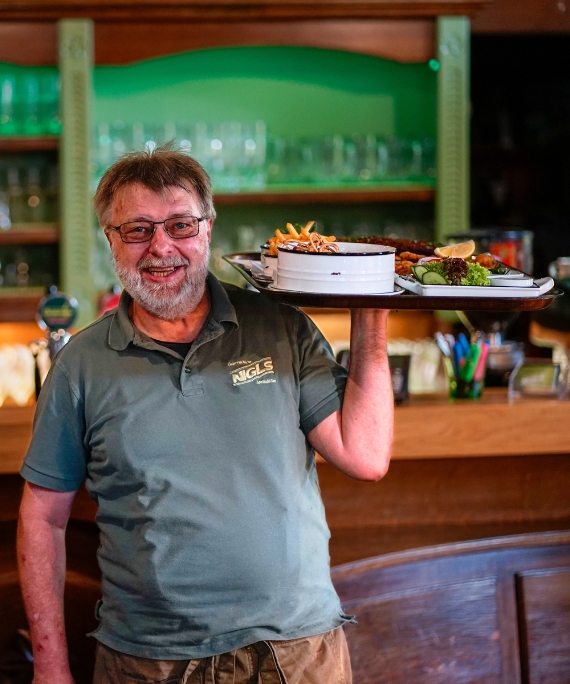 Kellner im Nigls lächelt freundlich in die Kamera und hält dabei ein Tablet voll mit Essen